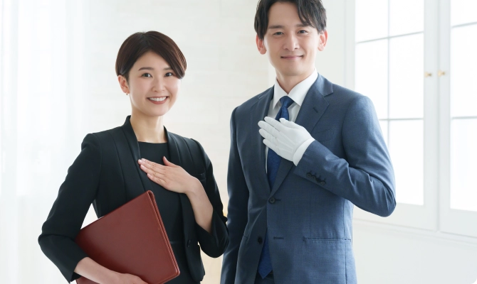 笑顔で迎えるビジネススーツ姿の男女社員。女性は胸に手を当て、男性は白い手袋をはめて胸に手を当てている。採用情報ページに掲載されており、プロフェッショナルで親しみやすい会社の雰囲気を表す。